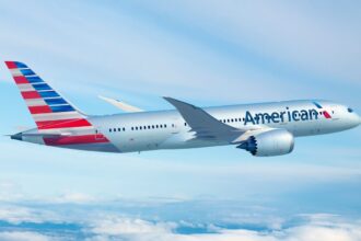 An red, white, and blue American Airlines jet flying in front of a cloudy sky.