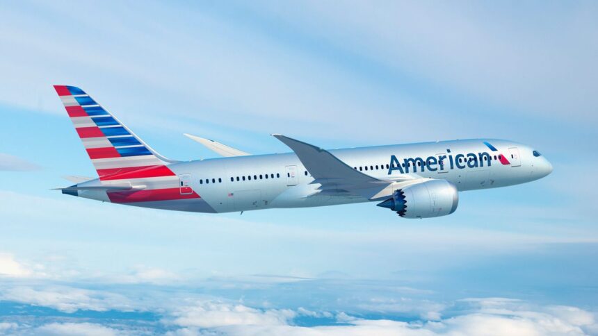 An red, white, and blue American Airlines jet flying in front of a cloudy sky.