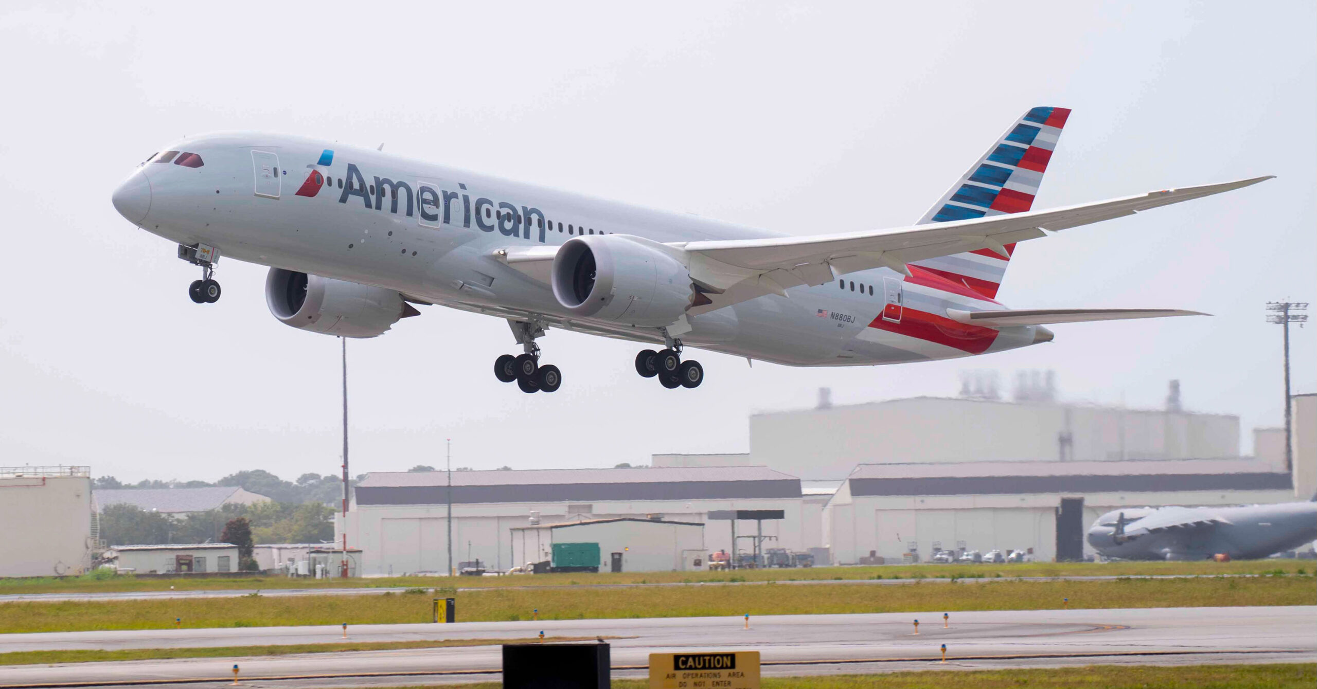 An American Airlines Boeing 787 landing on a runway in front of a grey sky, with an airport terminal behind it.