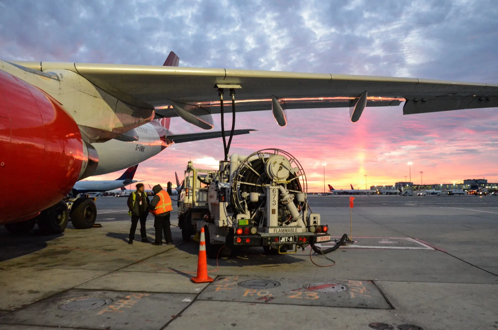 A red and white Airbus A330 being refuelled on an airport runway at sunset, by staff wearing hi-vis jackets.