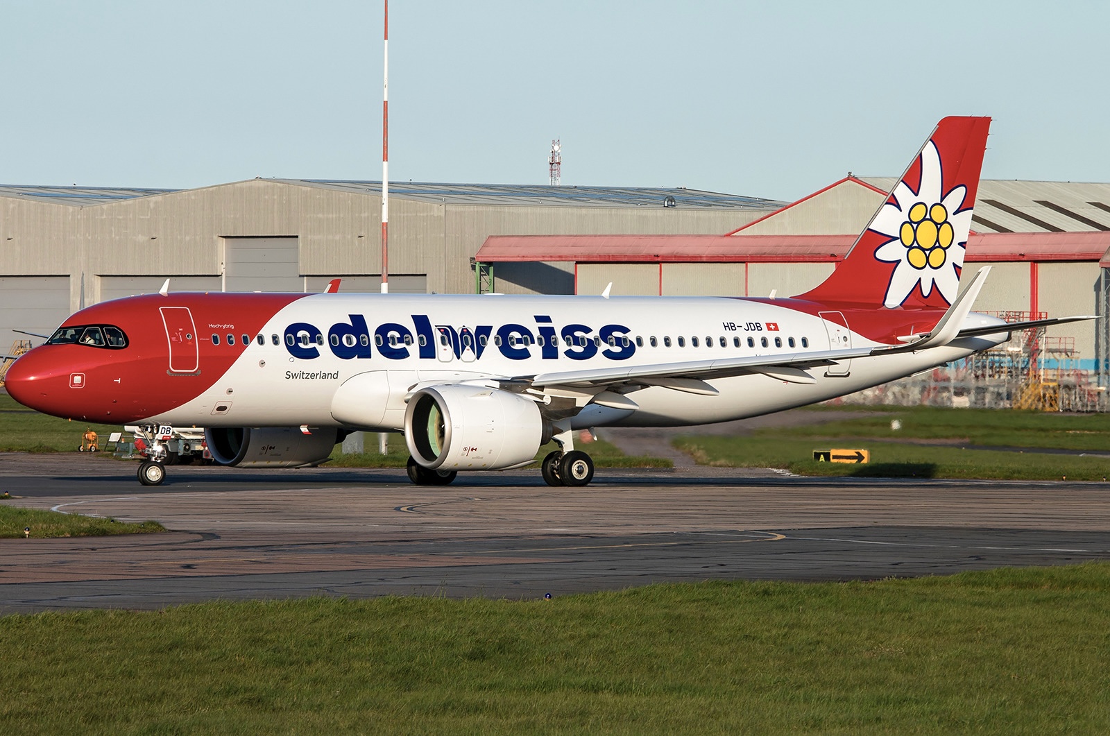 A red and white Edelweiss Airlines jet sat on a runway behind a verge of grass, and in front of an airport hanger.