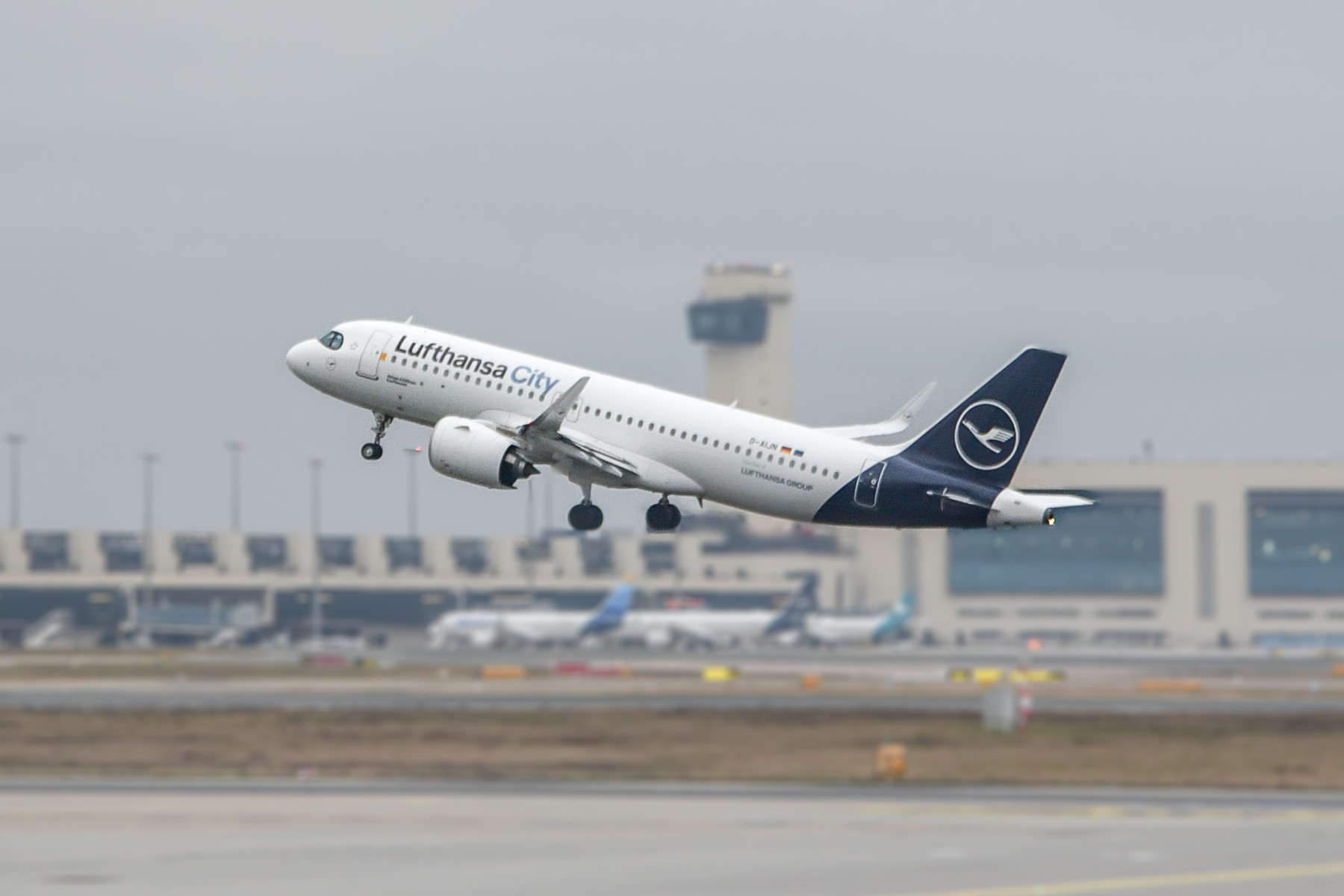 A blue and white Lufthansa City Airlines jet taking off from a runway in front of a dark grey sky.