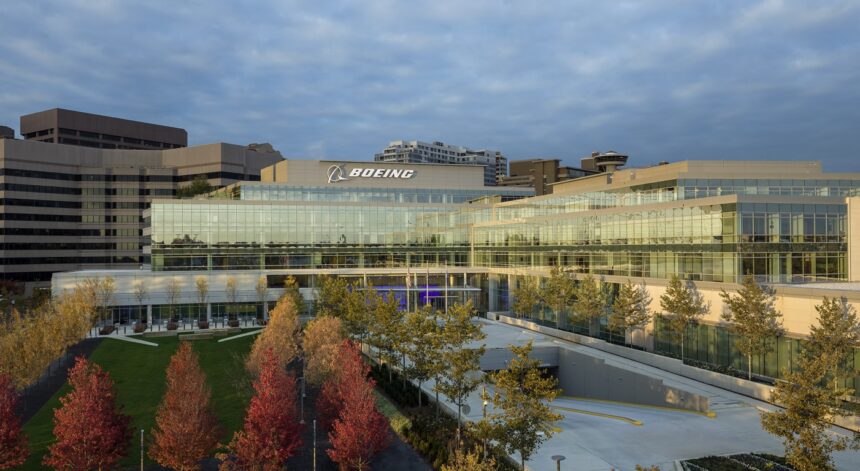 Boeing’s headquarters in Arlington, Virginia. A concrete and glass building is shown behind a clear lake and a small field surrounded by red trees.