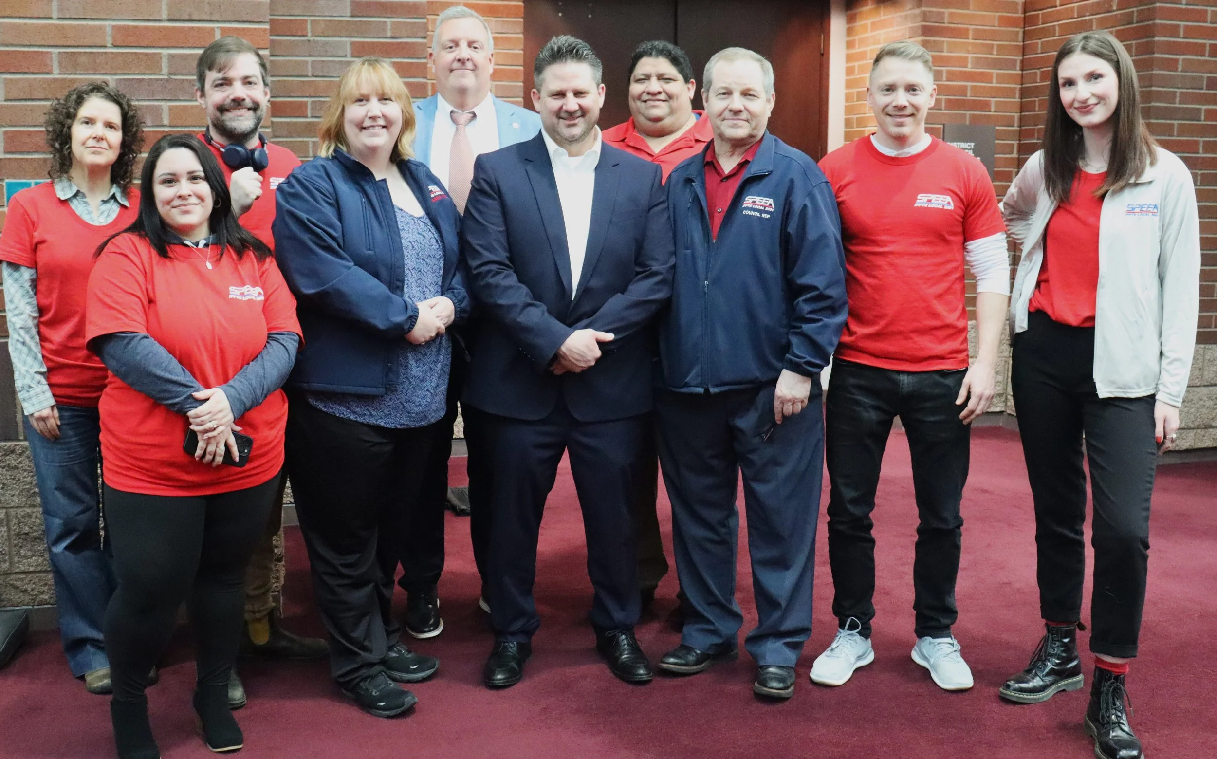 SPEEA members, many of who are dressed in red t-shirts with union’s logo. They are all stood indoors, on a purple carpet, in front of a brick wall.