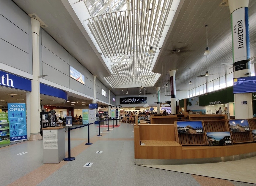 The inside of the terminal at Jersey Airport. The ceiling has glass so the day light peaks through. One can see half of a W.H Smith, a duty free, and a hot drinks area