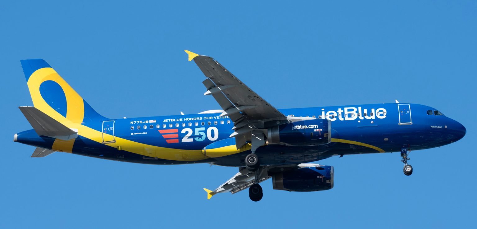 A Jetblue aircraft flying, blue fuselage with a yellow ribbon on the tail. 250 written in white, blue and red on the side of the aircraft. Blue sky in the background.
