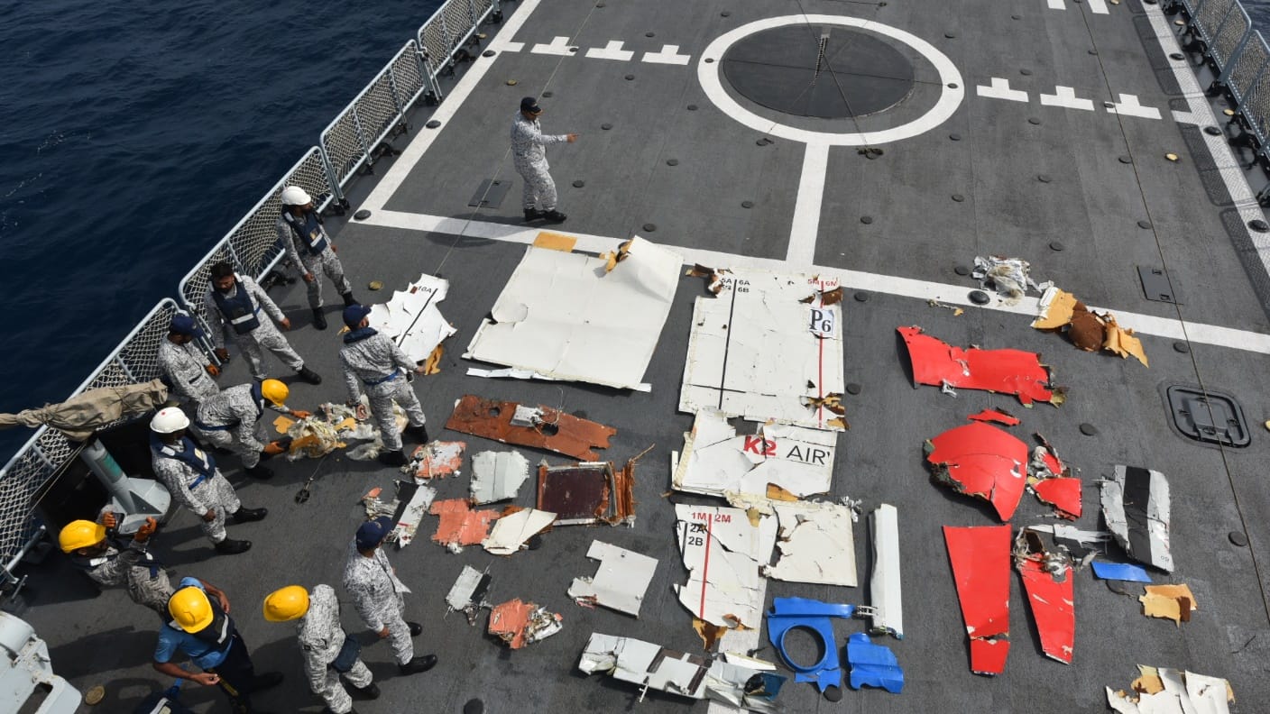 Aboard a large ship workers in hard hats and other such work gear surround the recovered wreckage of the Boeing 737 K2 Airways aircraft. Some parts are white with branding, others are red, some ae blue. The deck of the ship is grey with white line markings. The image is taken from above.