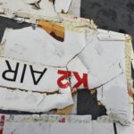 A piece of fuselage wreckage from the aircraft. It is jagged edged and whiteish in colour, with the red and black K2 airways branding visible. The background is the grey flooring of a large recovery vessel.