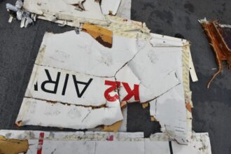 A piece of fuselage wreckage from the aircraft. It is jagged edged and whiteish in colour, with the red and black K2 airways branding visible. The background is the grey flooring of a large recovery vessel.