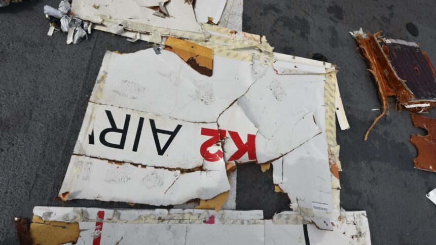 A piece of fuselage wreckage from the aircraft. It is jagged edged and whiteish in colour, with the red and black K2 airways branding visible. The background is the grey flooring of a large recovery vessel.