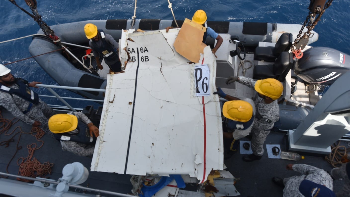 Another jagged edged broken piece of whiteish plane fuselage wreckage pictured from directly above. This piece is being held by 5 recovery staff in yellow hard hats. It is being transported from a smaller search boat (upper centre) to a larger recovery vessel(whole lower half of the image). The sea can be seen in the background along a strip at the top of the picture. 