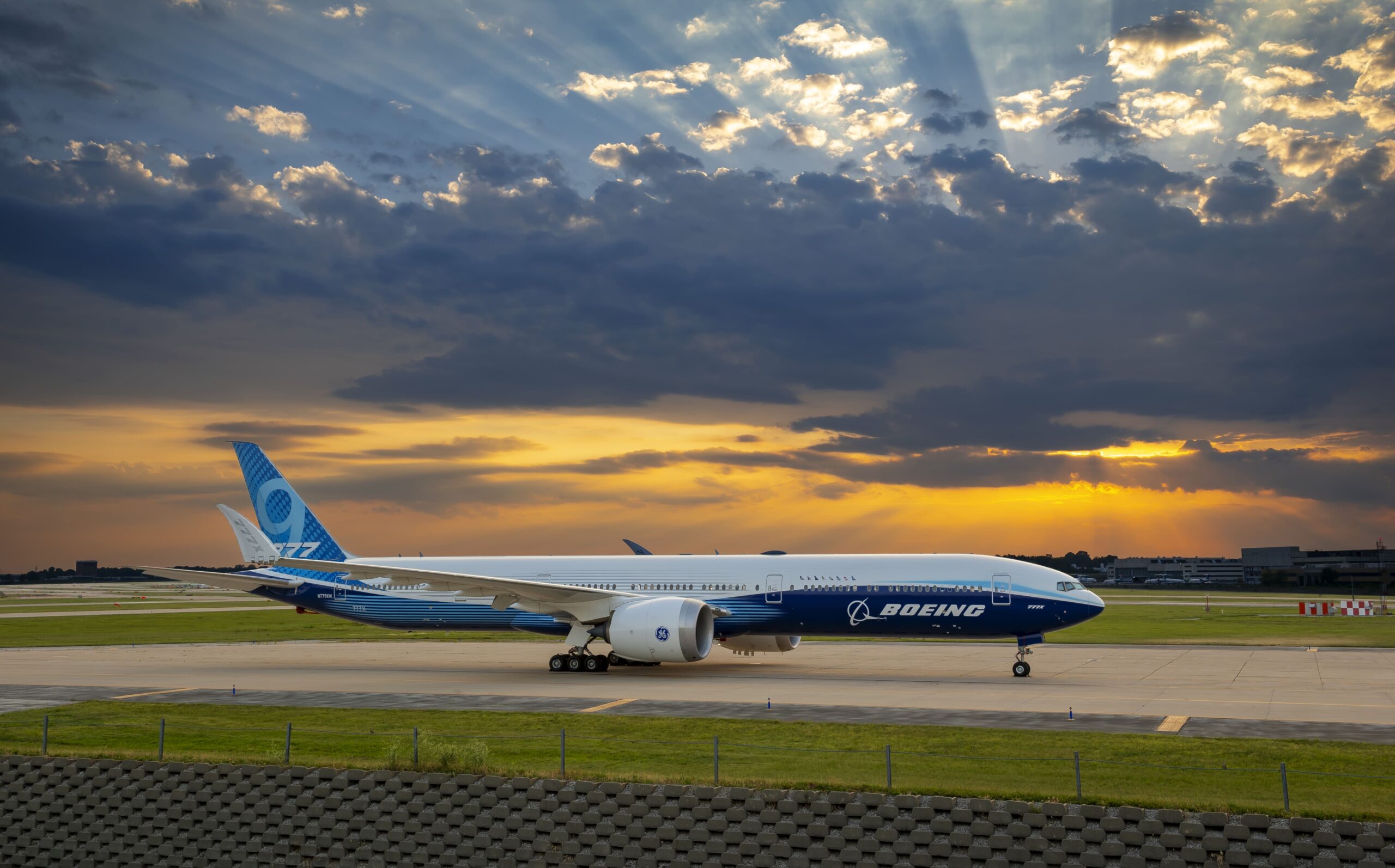 Boeing 777X aircraft in blue livery taxiing on runway at sunset.