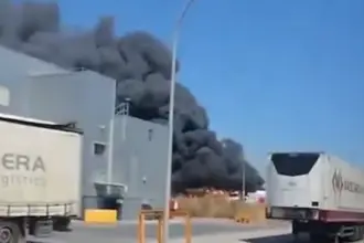 Smoke is seen billowing across Mallorca airport behind a building. Lorries pass on the road in front.