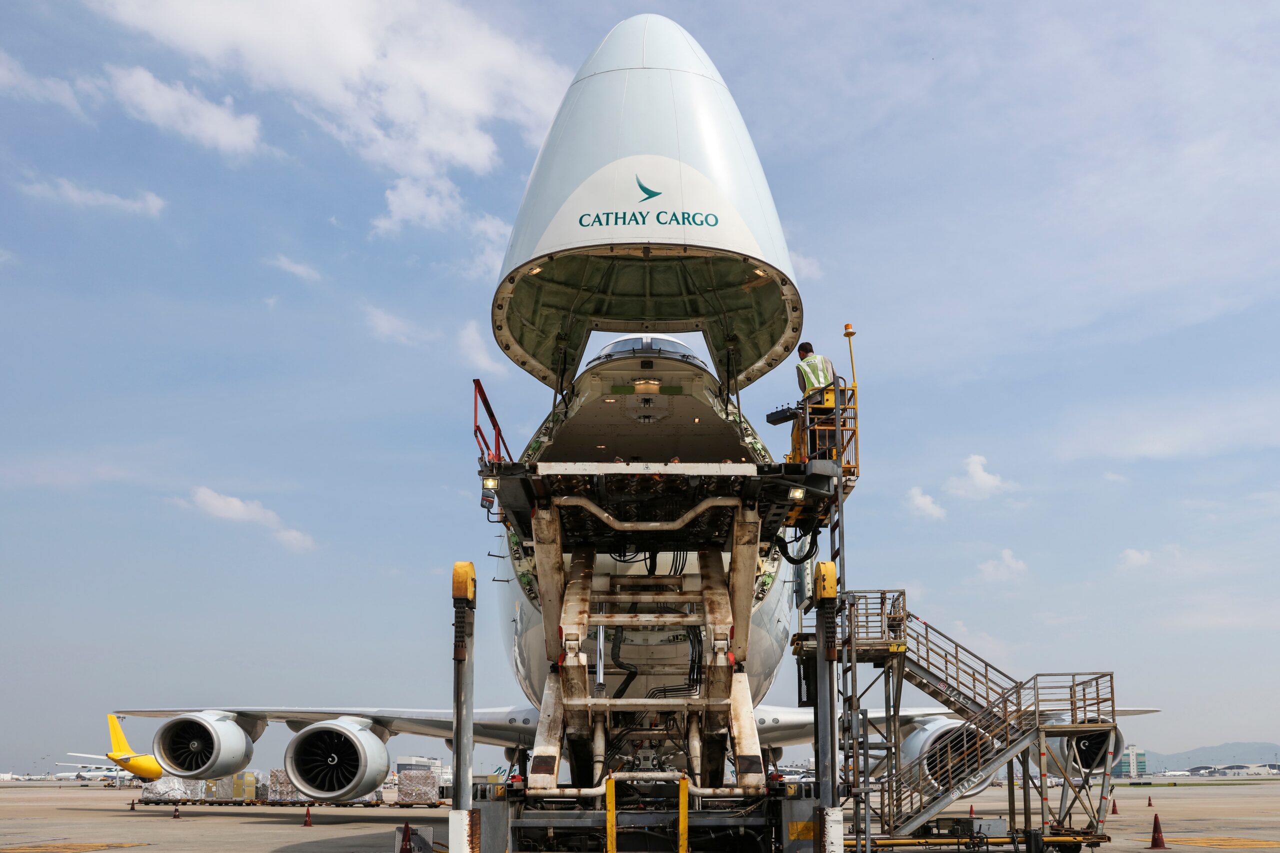 Image shows a grounded Cathay Cargo 747 freighter aircraft with its nose open and in the air and a worker at the top of steps nearby during the day