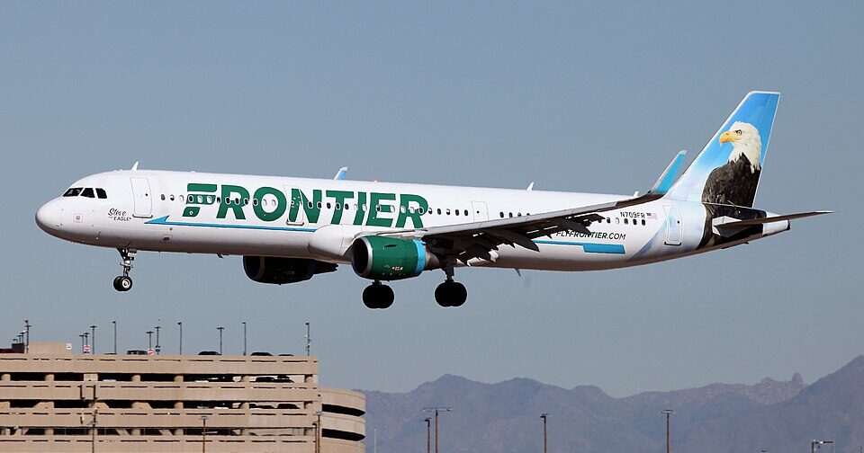 A Frontier Airlines Airbus A321 flying in to land, with its landing gear out. Classic white and green livery, with an eagle and blue background on the tail. Grey sky in the background.