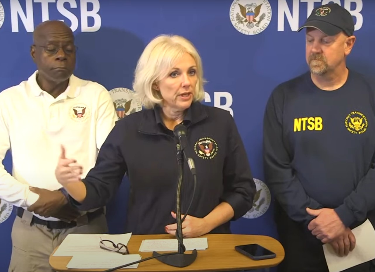 Jennifer Homendy, a middle aged blonde Caucasian female presenting person, stands at a podium with a microphone and documentation on it. Her mouth is partially open and she is gesticulating with her hand, implying she is giving a speech. She is wearing a navy blue zip through jacket with the NTSB logo on the right of her chest. Behind her on the left is a bald black male presenting person with glasses, and behind her on her right is a Caucasian male presenting person in a hat with a goatee beard. Both of these people also bear the NTSB logo on their clothing. 
