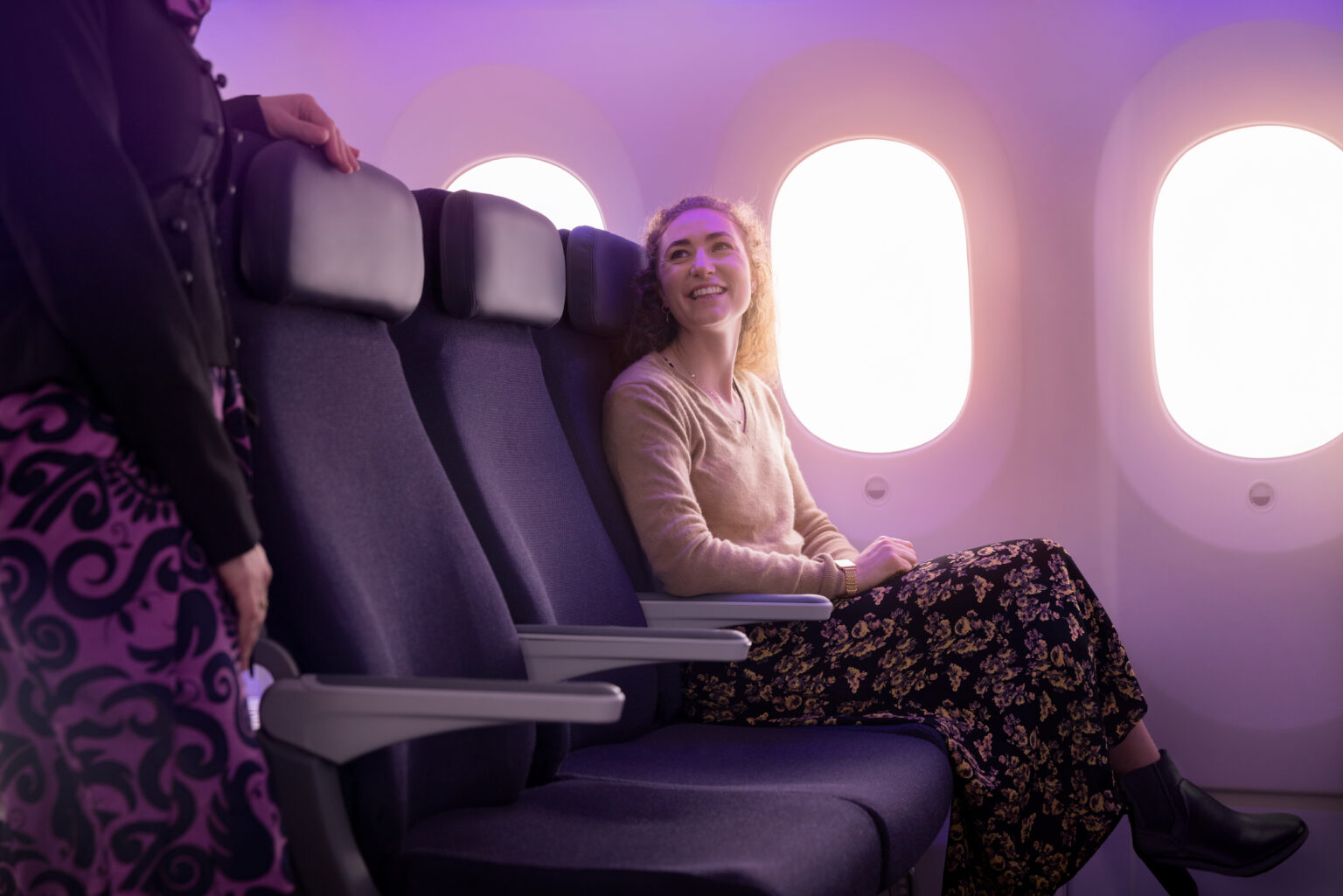 Passenger sat on the aeroplane window seat, laughing while talking to the Air hostess