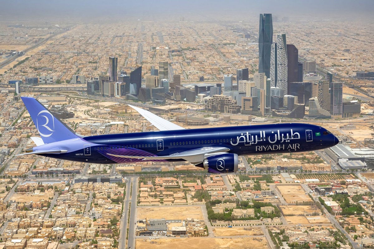 A Riyadh Air B787-9 flying over a city and several tall buildings. The livery is dark blue with a light blue tail. 
