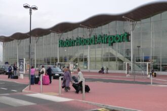 The outside front entrance to Doncaster Sheffield Airport (DSA) also known as Robin Hood Airport in Finningley. The picture was taken in 2006, the year after it opened. The building has a glass facade exterior wall, with 'Robin Hood Airport' in green lettering across it, beneath a scalloped style roof. The foreground of the image shows a tarmac road with a zebra crossing that people are crossing, as well as redbrick style pavement walkway. The sky appears grey and cloudy in the background.