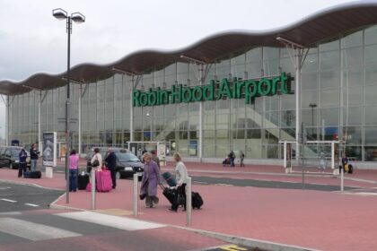 The outside front entrance to Doncaster Sheffield Airport (DSA) also known as Robin Hood Airport in Finningley. The picture was taken in 2006, the year after it opened. The building has a glass facade exterior wall, with 'Robin Hood Airport' in green lettering across it, beneath a scalloped style roof. The foreground of the image shows a tarmac road with a zebra crossing that people are crossing, as well as redbrick style pavement walkway. The sky appears grey and cloudy in the background.