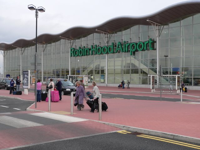 The outside front entrance to Doncaster Sheffield Airport (DSA) also known as Robin Hood Airport in Finningley. The picture was taken in 2006, the year after it opened. The building has a glass facade exterior wall, with 'Robin Hood Airport' in green lettering across it, beneath a scalloped style roof. The foreground of the image shows a tarmac road with a zebra crossing that people are crossing, as well as redbrick style pavement walkway. The sky appears grey and cloudy in the background.