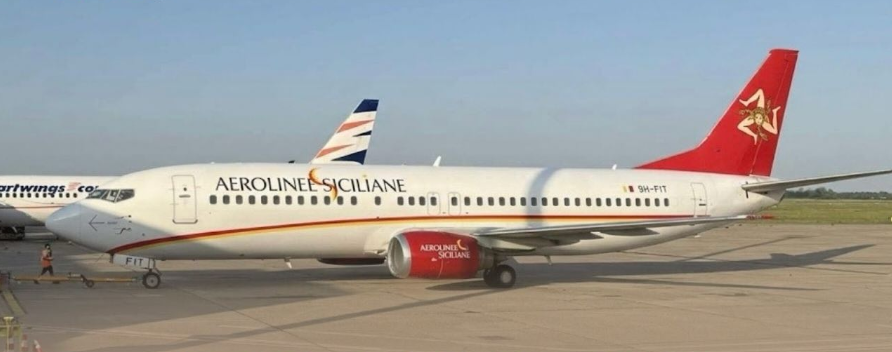 An Aerolinee Siciliane aircraft sitting in the apron. White, blue, red and yellow livery. Blue sky in the background, and grass visible on the left side.