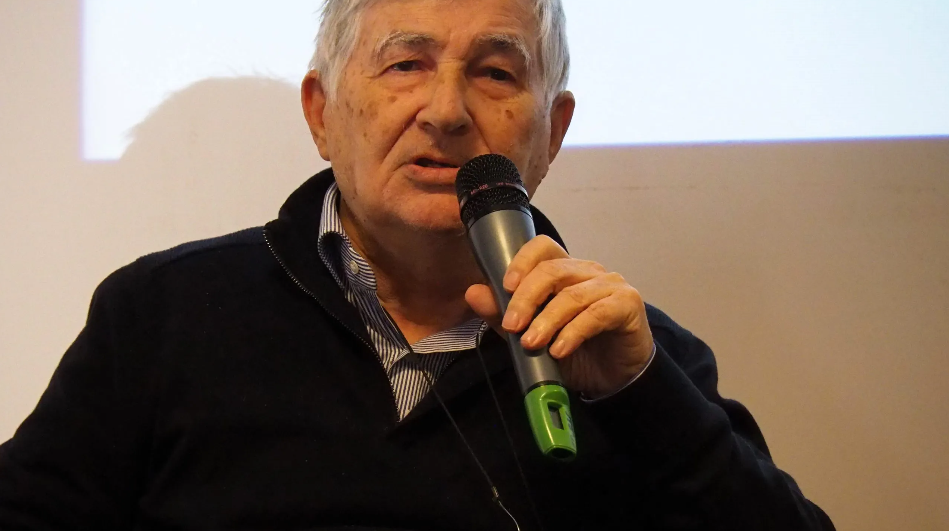 Luigi Crispino, wearing a button up shirt and a black jacket, sat down holding a microphone, talking. Sat in front of a projector screen.