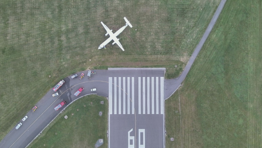 A white plane seen on the grass past the runway, with emergency vehicles, including fire, at the scene toward the bottom left.