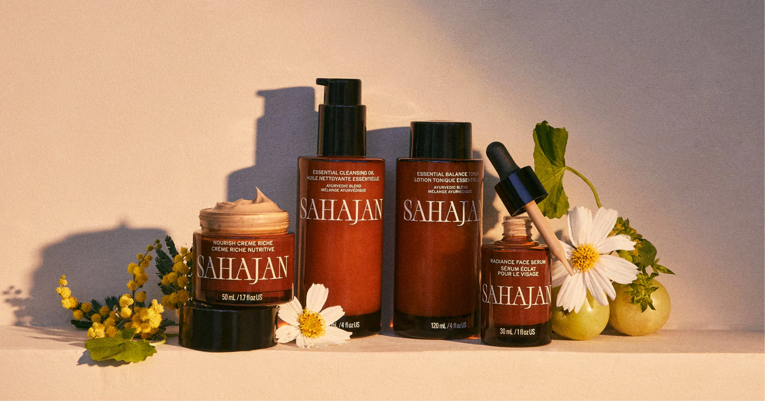 Sahajan products lined up alongside flowers and leaves in brown bottles, featuring creams, eye drops and cleansers.