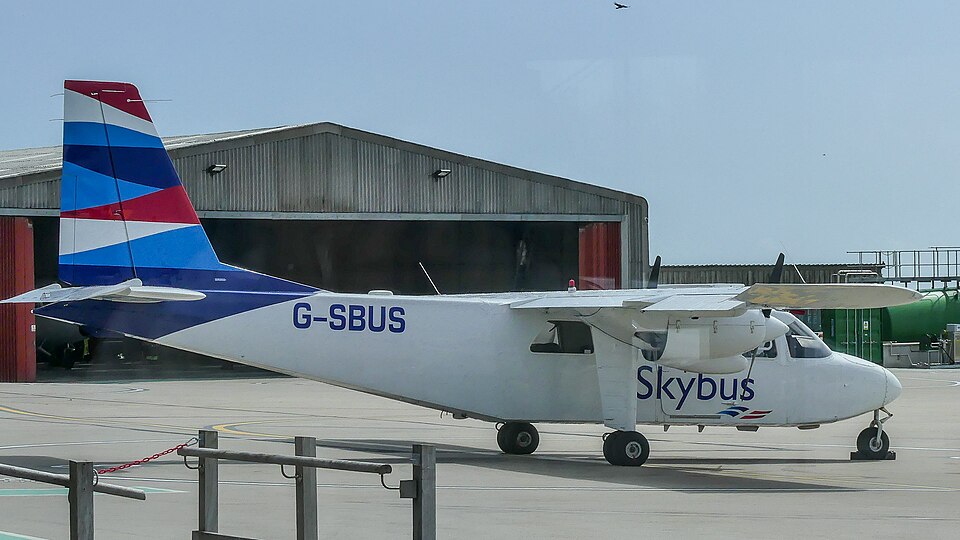 A Skybus aircraft on the tarmac before takeoff. Classic white, red and blue livery. Registration G-SBUS. Blue sky and building in the background.