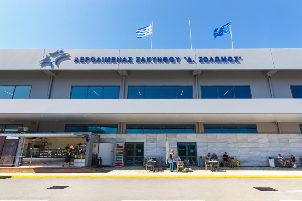 An image of the front of Zakynthos Airport with the airport sign and greek and EU flags as well as people waiting outside the front of the building. 