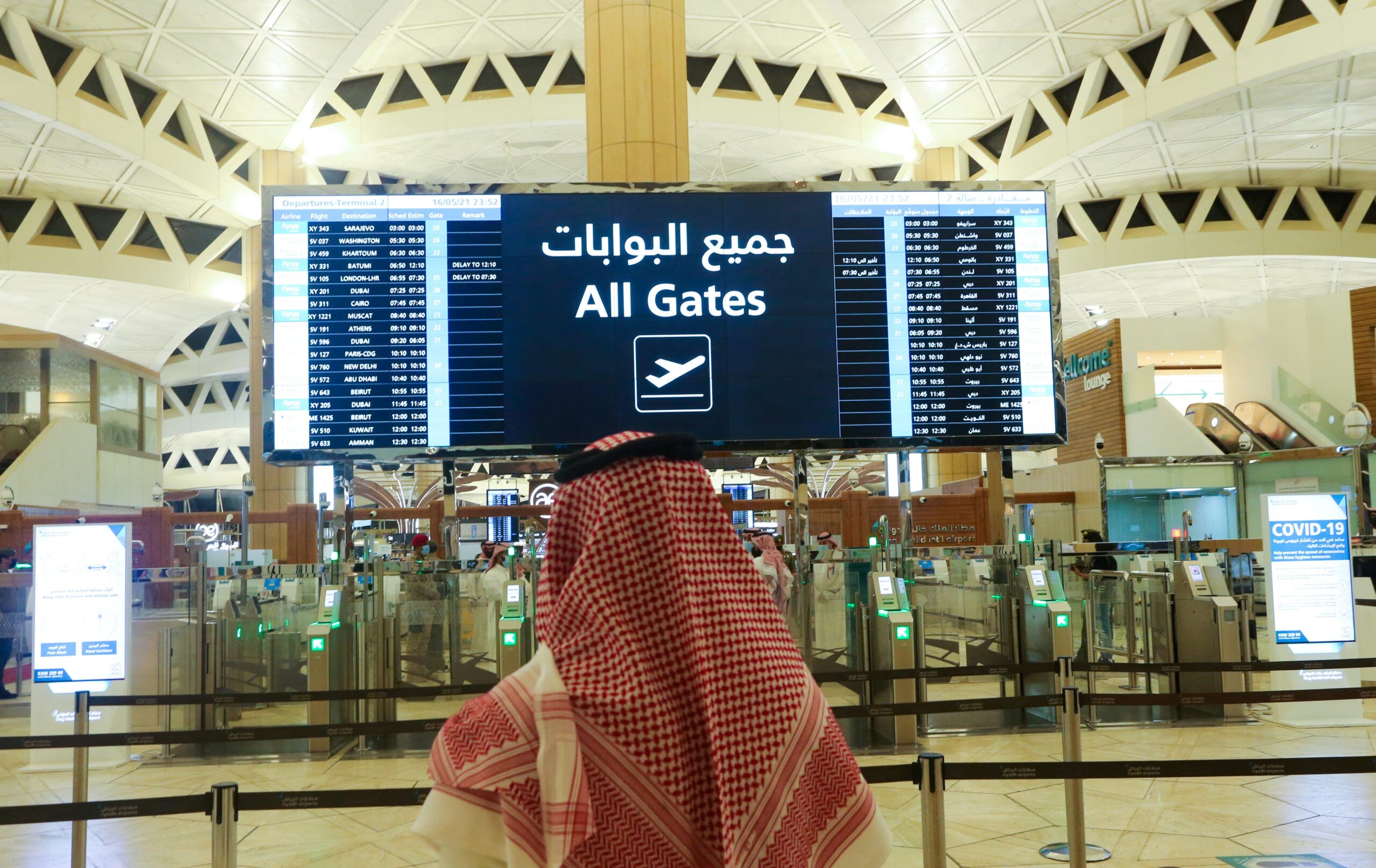 Man standing in front of departures board at an airport in Saudi Arabia with the gates in front, wearing traditional clothing