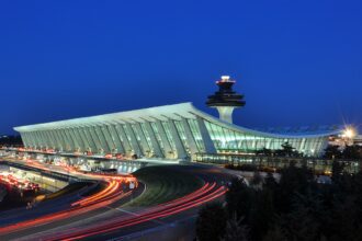 Washington DC Dulles International Airport at night. The airport is covered in glass and the control tower is seen behind it. There are also departure and arrival pick up and drop off spots visible.