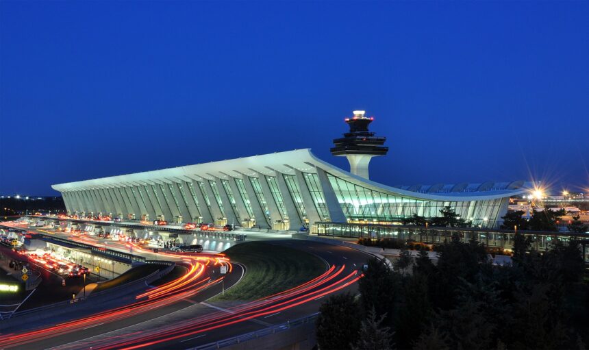Washington DC Dulles International Airport at night. The airport is covered in glass and the control tower is seen behind it. There are also departure and arrival pick up and drop off spots visible.