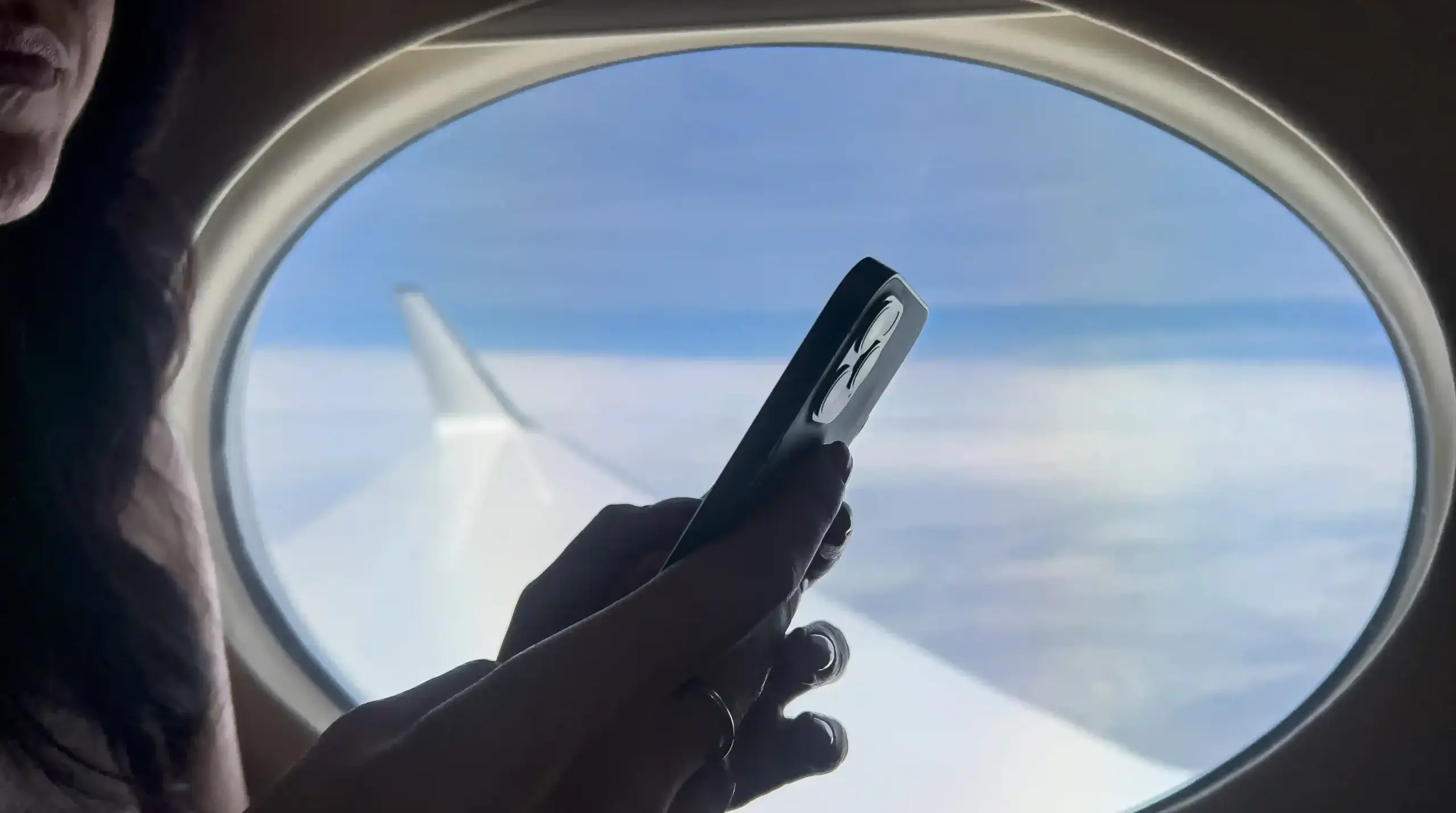 A women holding her phone up on a plane in front of a window. Outside the window is the plane's wing and a blue sky in the background.