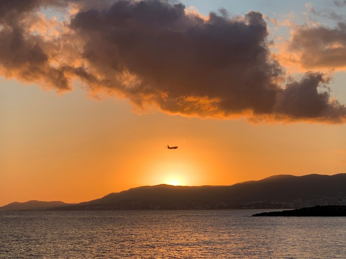 A plane flying over the horizon of a Mallorca sea with an orange sunset, clouds and a hill in the background