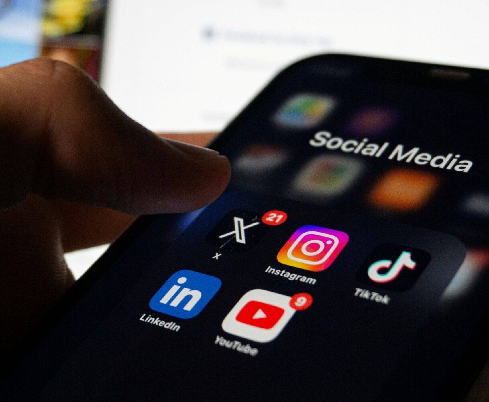 A zoomed in, stylistically blurred image of a phone screen displaying social media app icons. The phone is angled from the lower bottom left corner to upper centre right of the image. A Caucasian thumb can be see hovering over the app icon for X. 