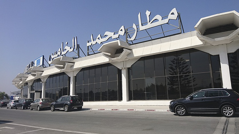 The front entrance of Mohammed V International Airport in Casablanca with a large sign on top of the building with the airport's name. Some dark coloured cars are parked in front of the airport.