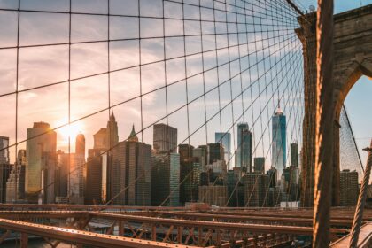 A city skyline at sunset viewed through the cables of a suspension bridge, with skyscrapers illuminated by warm sunlight and the bridge structure framing the foreground