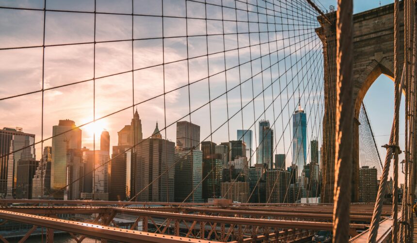 A city skyline at sunset viewed through the cables of a suspension bridge, with skyscrapers illuminated by warm sunlight and the bridge structure framing the foreground