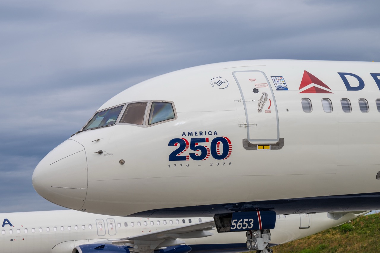 A close up shot of Delta Air Lines nose of the plane. "America 250" is written in blue and red, against a white plane. Blue, cloudy sky in the background.