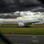 A side view of an Emirates aircraft on the Birmingham Airport tarmac with grass surrounding it and a dark cloudy sky.