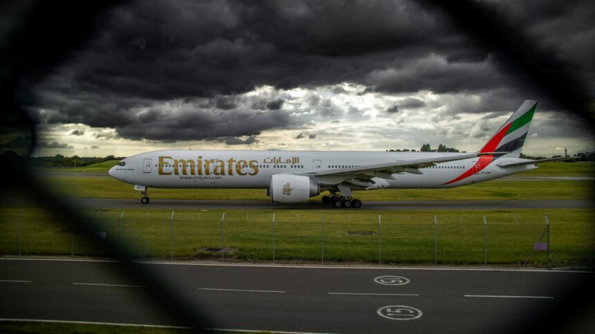 A side view of an Emirates aircraft on the Birmingham Airport tarmac with grass surrounding it and a dark cloudy sky.