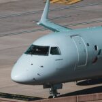 The nose of an Embraer aircraft is pictured from quite close while the aircraft is grounded. The aircraft's nose and the cockpit is white with 'Embraer' branding in blue beneath the cockpit windscreen. The background shows the grey-ish brown concrete of an airport taxiway or hangar or runway.