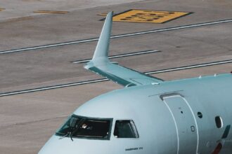 The nose of an Embraer aircraft is pictured from quite close while the aircraft is grounded. The aircraft's nose and the cockpit is white with 'Embraer' branding in blue beneath the cockpit windscreen. The background shows the grey-ish brown concrete of an airport taxiway or hangar or runway.