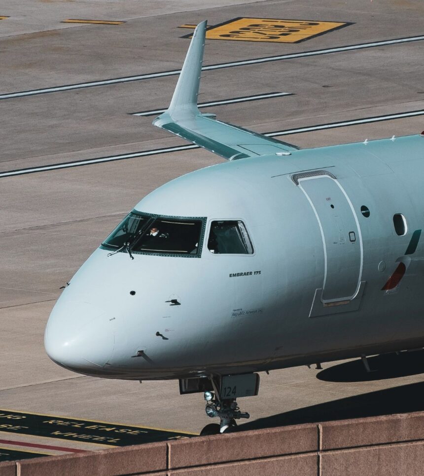 The nose of an Embraer aircraft is pictured from quite close while the aircraft is grounded. The aircraft's nose and the cockpit is white with 'Embraer' branding in blue beneath the cockpit windscreen. The background shows the grey-ish brown concrete of an airport taxiway or hangar or runway.
