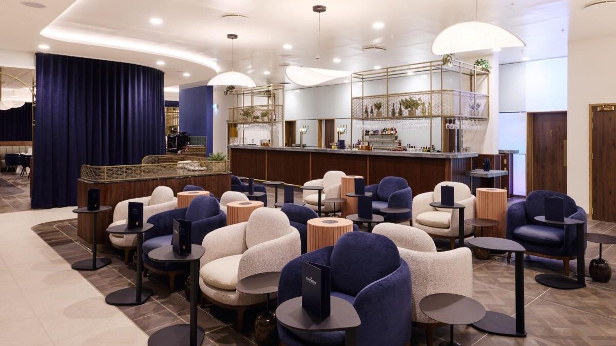Interior of the new lounges at MAN. Blue and white cushioned chairs around small circular tables. Bright lights handing down from the ceiling, with a bar in the background.