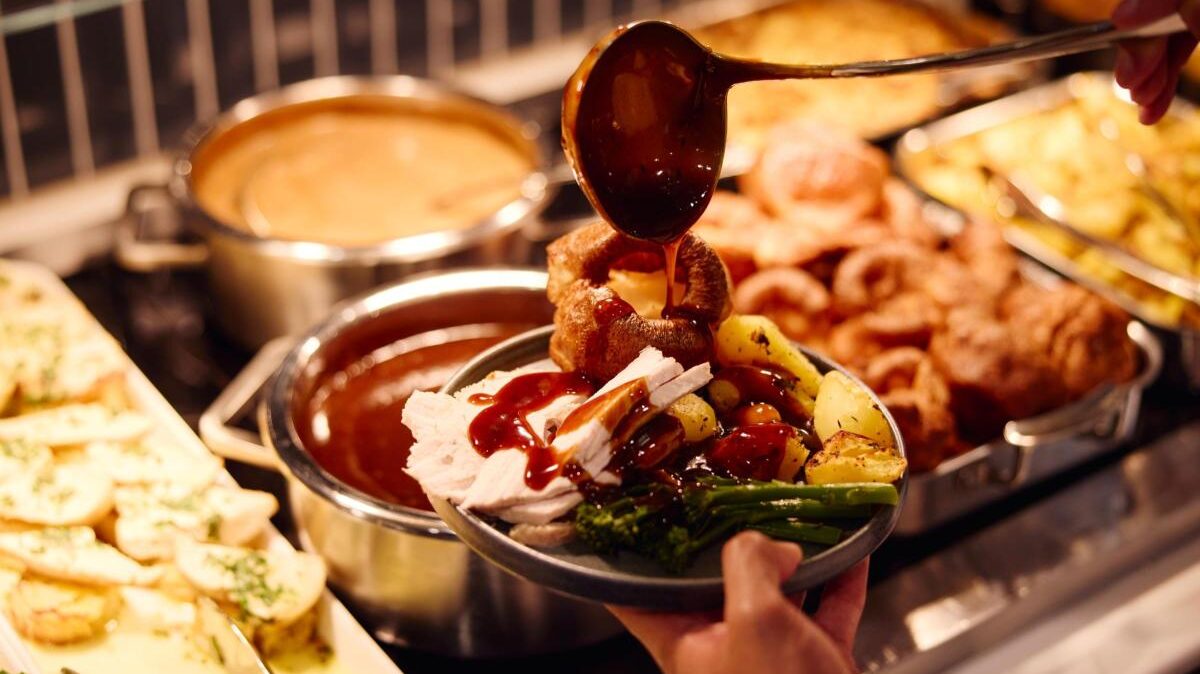 A carvery at MAN. A plate filled with food, while someone pours gravy over it. Pots of food seen in the background under lighting.