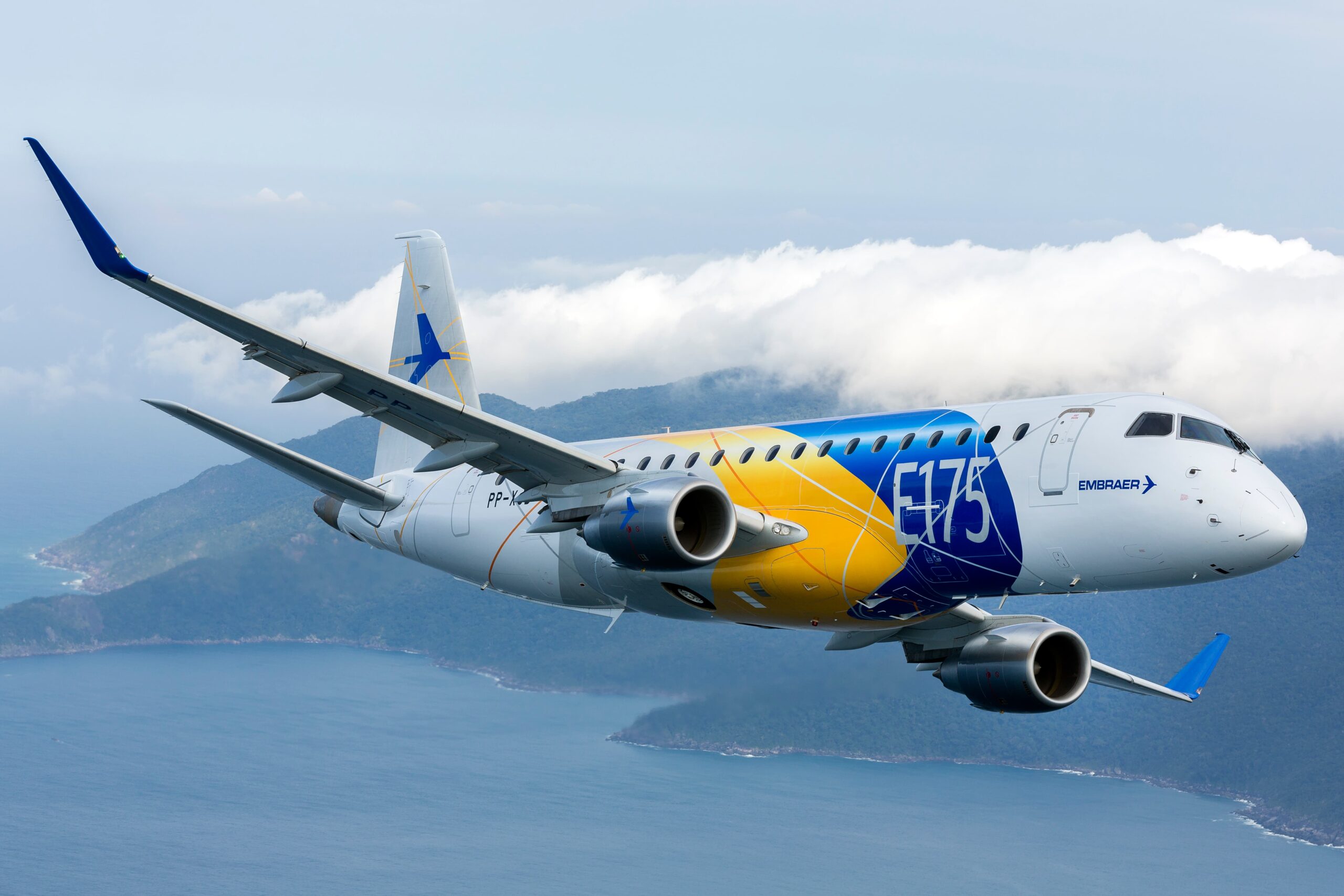 Embraer E175 side view flying above the sea with an island and clouds in the background. Aircraft is branded in yellow and blue colours