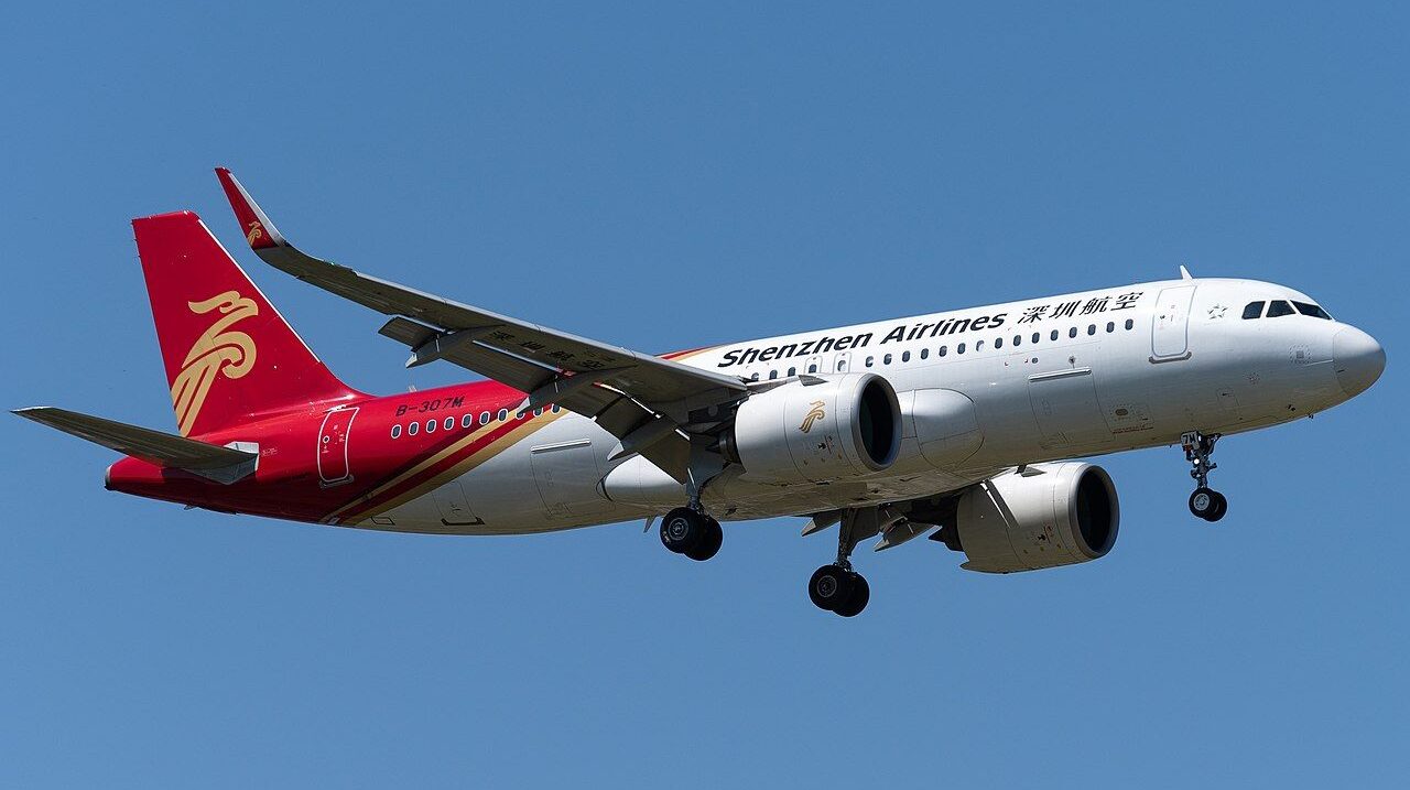 A Shenzhen Airlines Airbus A320 flying in the blue sky toward the right side of the frame. Classic white, red and yellow livery, with its landing gear out.